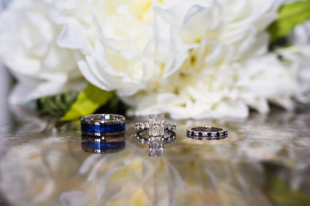 Three wedding rings are arranged on a reflective surface in front of softly blurred white flowers. A dark metal groom’s band with a vivid blue center sits beside a diamond engagement ring with a rectangular center stone and a slender wedding band set with alternating blue and clear stones.