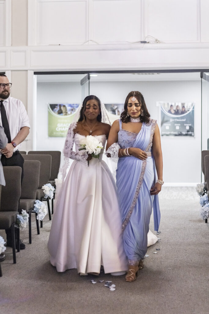An emotional bride walks down the aisle of a New Jersey Wedding Chapel arm in arm with her mother, wearing a pale blue sari embellished with silver details. The bride holds a bouquet of white flowers and wears a long lace veil as the pair walk between rows of gray chairs decorated with pale blue and white flowers.