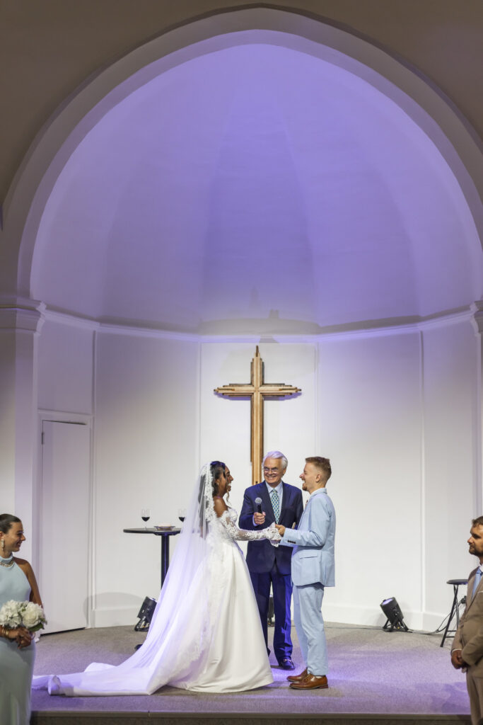 A bride and groom face one another and hold hands while exchanging vows beneath the tall arched ceiling of a New Jersey Wedding Chapel. An officiant stands smiling between them with a microphone, while a large wooden cross is centered above the couple and purple light softly illuminates the church ceiling.