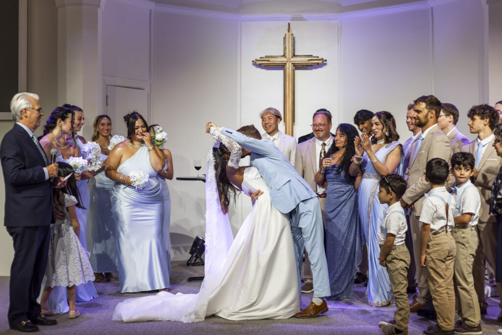 A groom dramatically dips and kisses his bride at the altar of a New Jersey Wedding Chapel as their family and wedding party laugh, clap and watch around them. The bride’s long white veil and full gown sweep toward the floor beneath a large wooden cross.