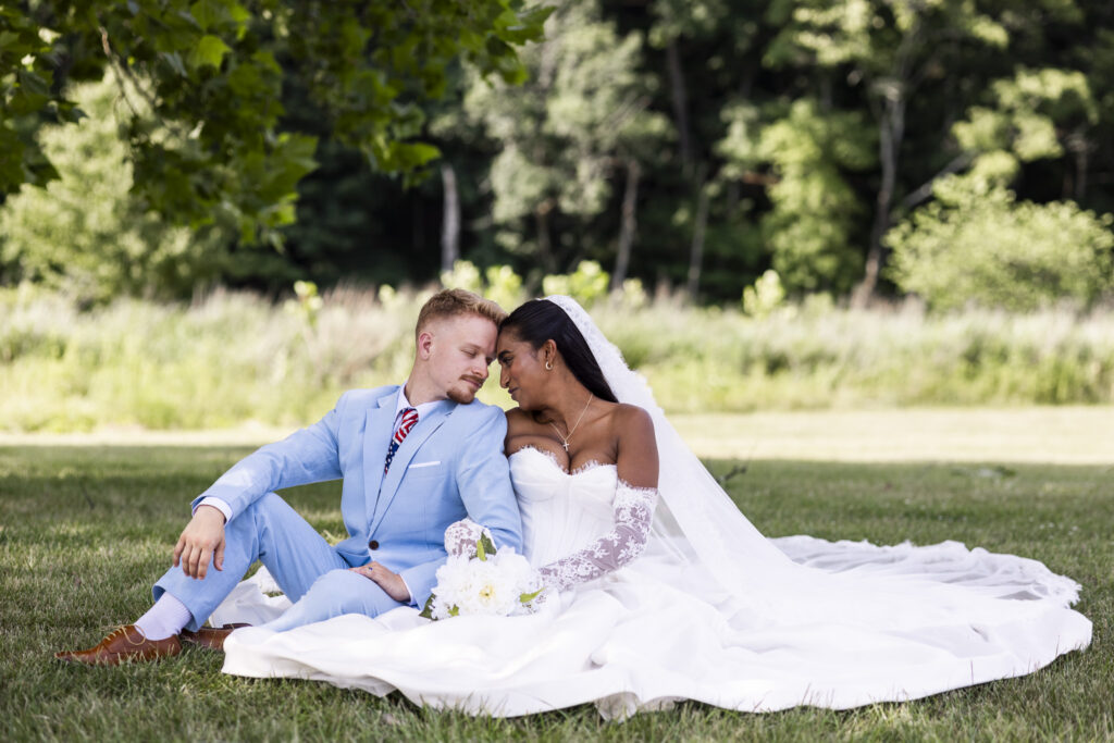 A bride and groom sit closely together on the grass beneath the shade of a tree during their wedding portraits. The groom, dressed in a pale blue suit, leans his forehead toward the bride as she rests beside him in her white ball gown and lace sleeves. Her long veil and gown spread dramatically across the grass around them.