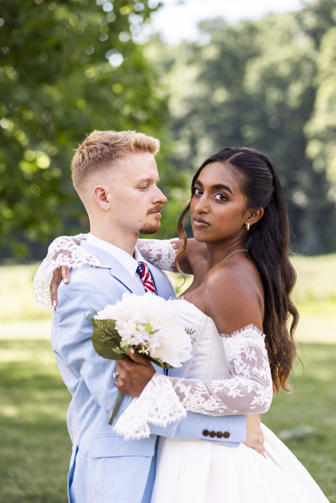 A close outdoor wedding portrait shows a bride and groom embracing against a softly blurred green landscape after their New Jersey Wedding Chapel ceremony. The bride holds a white bouquet and looks directly toward the camera while wrapping one lace-covered arm around the groom, who wears a pale blue suit and patriotic red, white and blue tie.