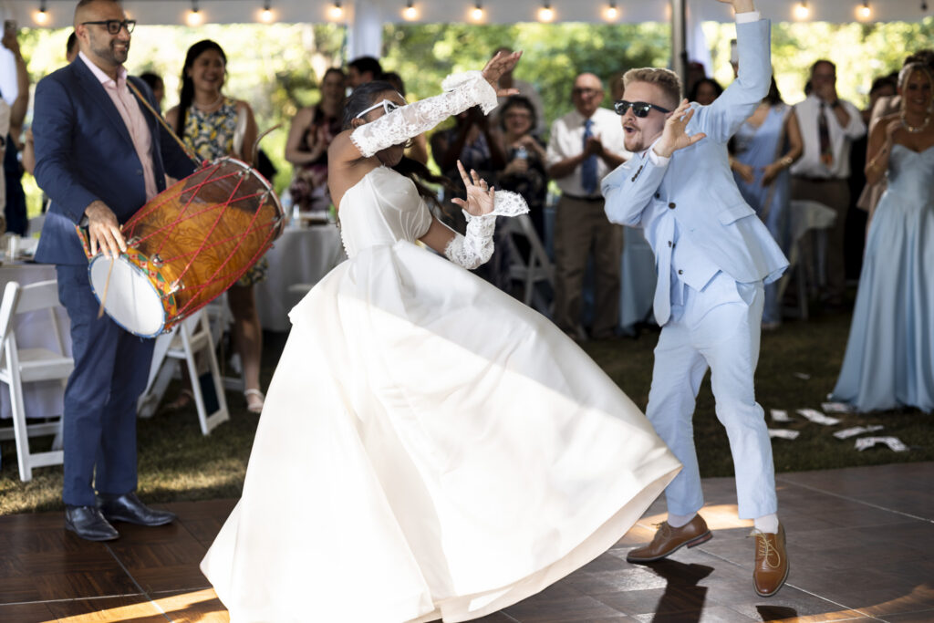 A bride and groom dance energetically beneath a white tent during their backyard wedding reception as a musician plays a large traditional drum beside them. The bride’s full skirt swirls across the dance floor while cheering guests watch from behind.