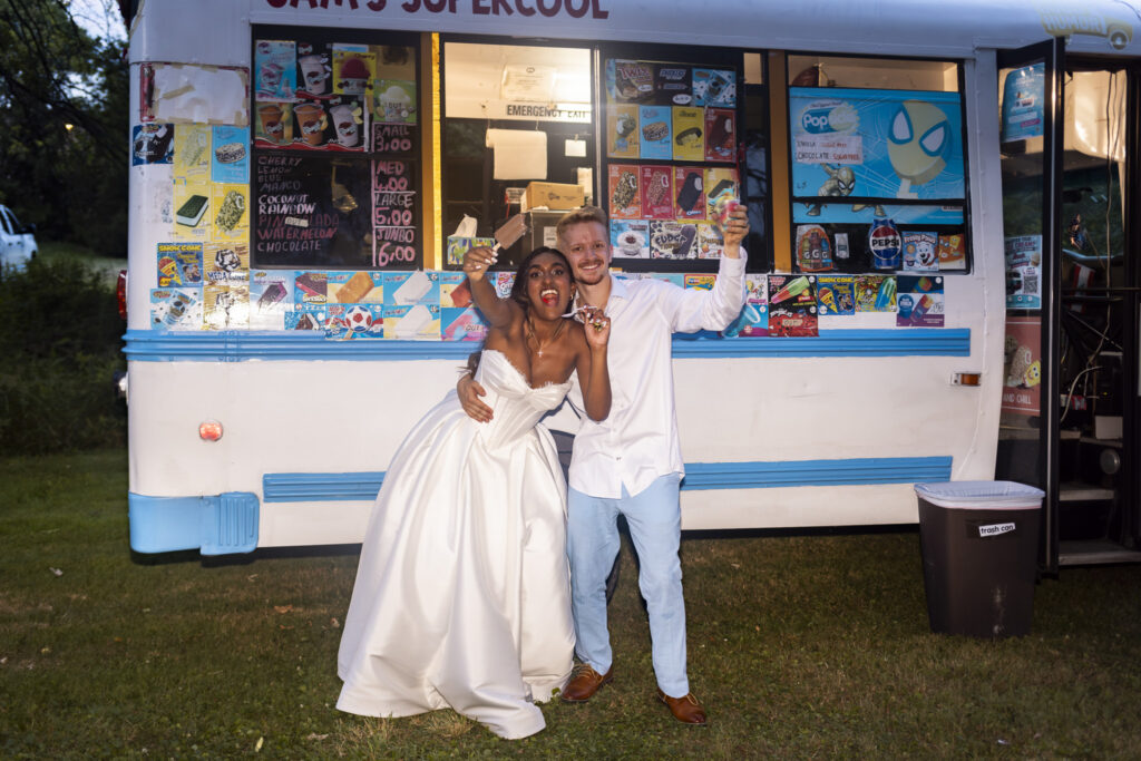 A bride and groom enthusiastically hold up frozen treats while posing in front of a white and blue ice cream truck at their backyard wedding reception. The bride wears her white wedding gown while the groom has removed his jacket and wears a white shirt with pale blue trousers. The truck’s colorful menu of ice cream bars and frozen treats fills the background.