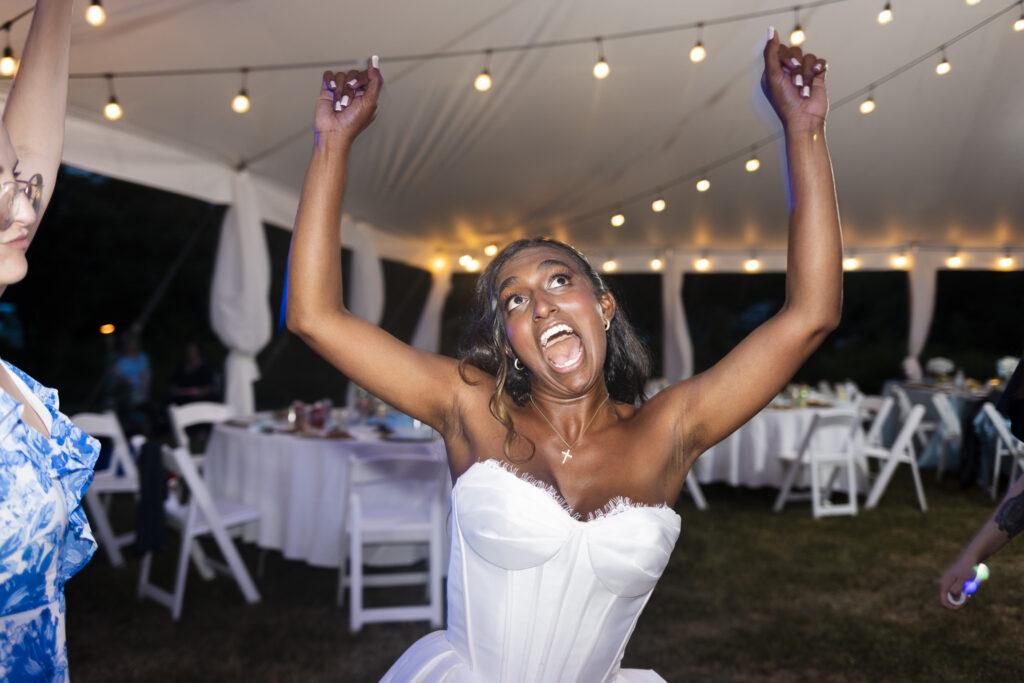 A bride throws both arms high above her head while dancing beneath a white reception tent at night. Her mouth is open in an exuberant expression as she celebrates beneath strands of glowing café lights, with white reception tables and chairs visible behind her.