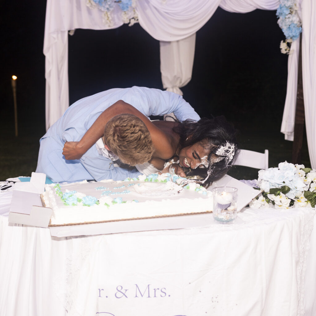 A bride and groom laugh with their arms around one another immediately after a playful wedding cake smash. White frosting covers one side of the bride’s face, hair and bodice, while cake is also smeared across the groom’s face. A large partially smashed sheet cake sits on the table in front of them beneath a white-and-blue floral reception backdrop.