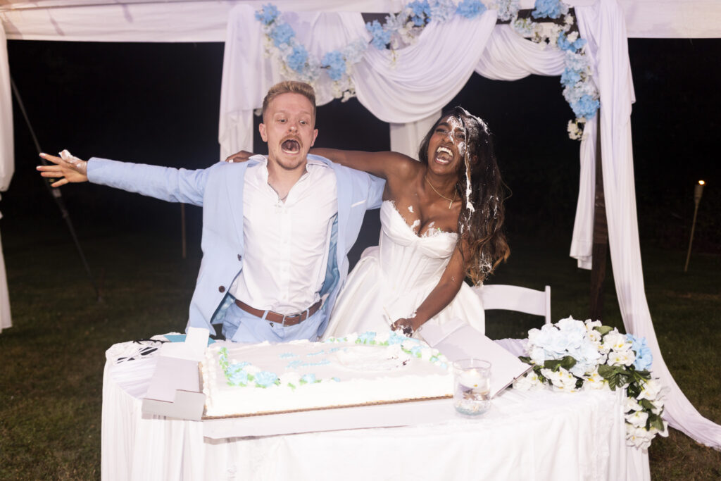 A bride and groom laugh together behind their wedding cake during a nighttime backyard wedding reception, as the bride pushes her groom towards the cake during a playful cake-smashing moment. Pale blue flowers and flowing white fabric decorate the backdrop behind them.