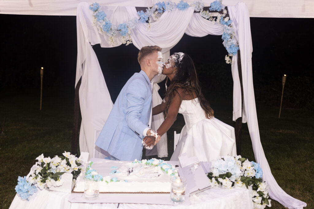 A newlywed couple covered in wedding cake frosting leans across their sweetheart table to kiss during their backyard wedding reception. The partially smashed white cake sits between arrangements of pale blue and white flowers beneath a draped reception backdrop.