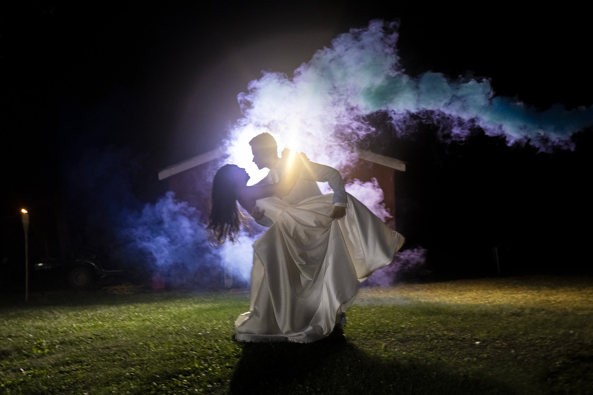 A bride and groom are silhouetted against a bright backlight while he dips her for a dramatic nighttime wedding portrait. Clouds of blue, purple and turquoise smoke billow around the couple, creating a colorful ending to their New Jersey Wedding Chapel celebration.