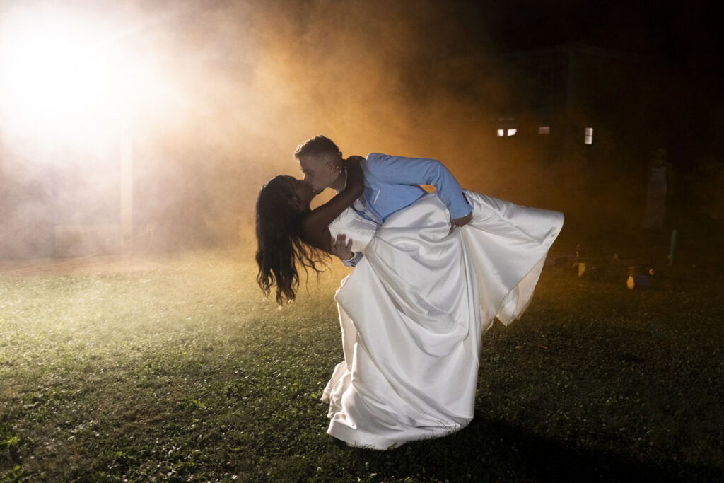 A groom dips and kisses his bride outdoors at night during their backyard wedding reception, surrounded by glowing golden light and atmospheric smoke. Her full white wedding gown billows outward as he supports her in the dramatic portrait.