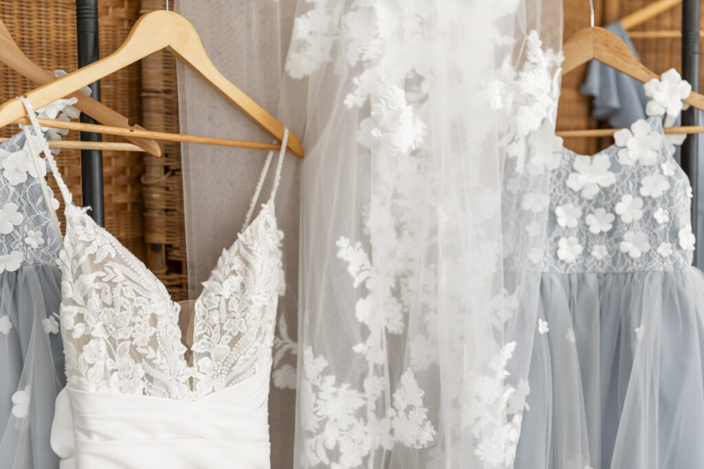 White lace wedding gown with a plunging neckline and floral appliqué hangs beside dusty blue flower girl dresses, capturing the soft blue and white palette of a south jersey wedding.