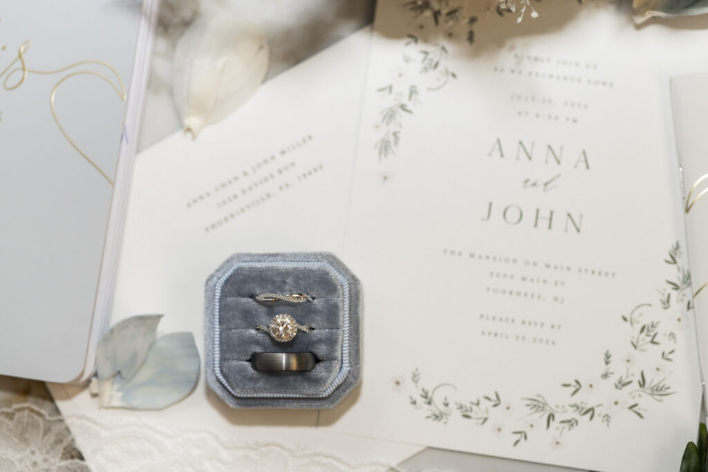 Three wedding rings rest inside a blue velvet ring box on top of a floral wedding invitation, surrounded by delicate stationery and detail pieces from a south jersey wedding.