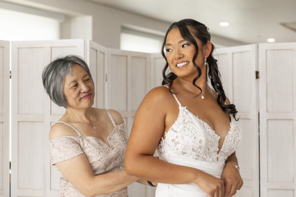 A mother smiles as she helps fasten the back of her daughter’s white lace wedding dress during the getting-ready portion of a south jersey wedding.