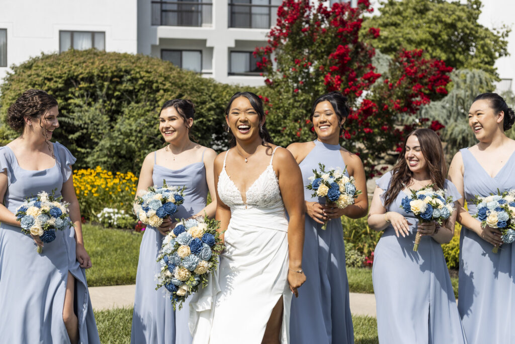 Bride laughs with five members of her wedding party in dusty blue dresses, each holding coordinating blue and ivory bouquets in the sunny gardens during a south jersey wedding.