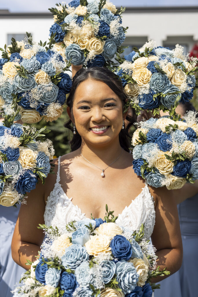 Bride smiles at the camera while holding a blue and ivory bouquet as matching bouquets held around her frame her face during a playful south jersey wedding portrait.