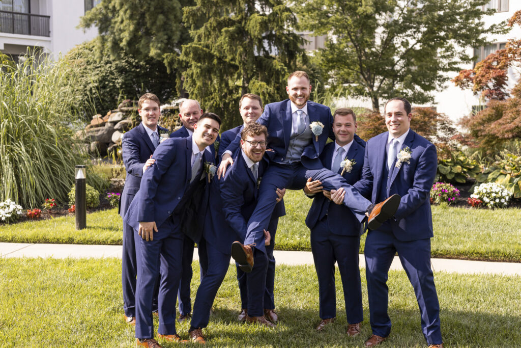 Groom in a blue suit is lifted horizontally by his groomsmen as the group laughs together outside among the landscaped gardens during a south jersey wedding.