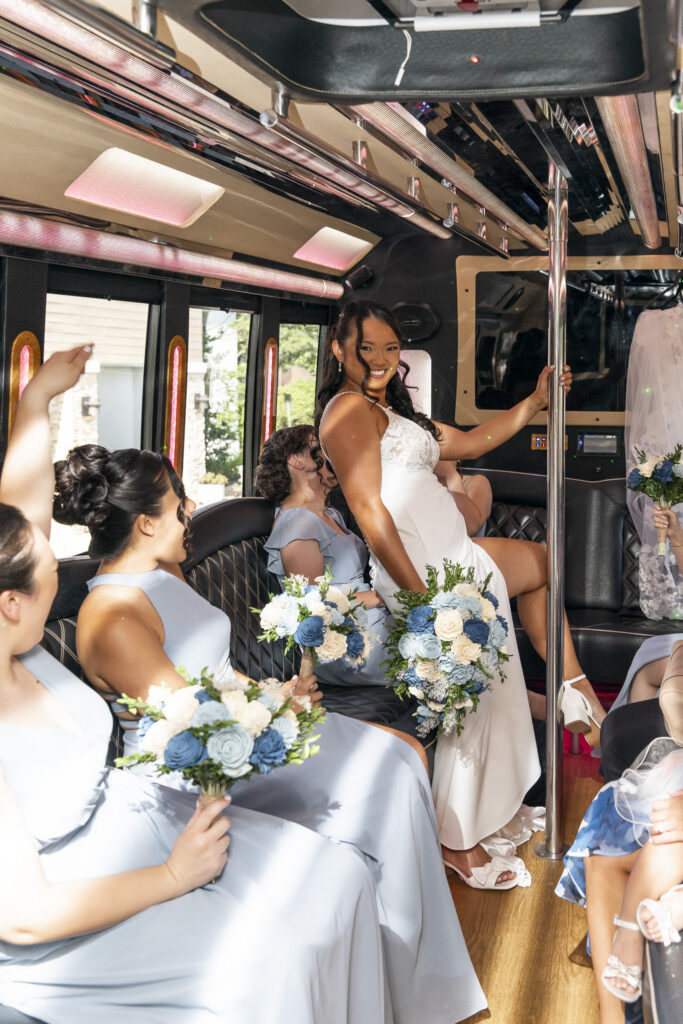 Bride playfully poses around a pole inside a party bus while her wedding party laughs from their seats, holding coordinating blue and ivory bouquets on the way to a south jersey wedding.