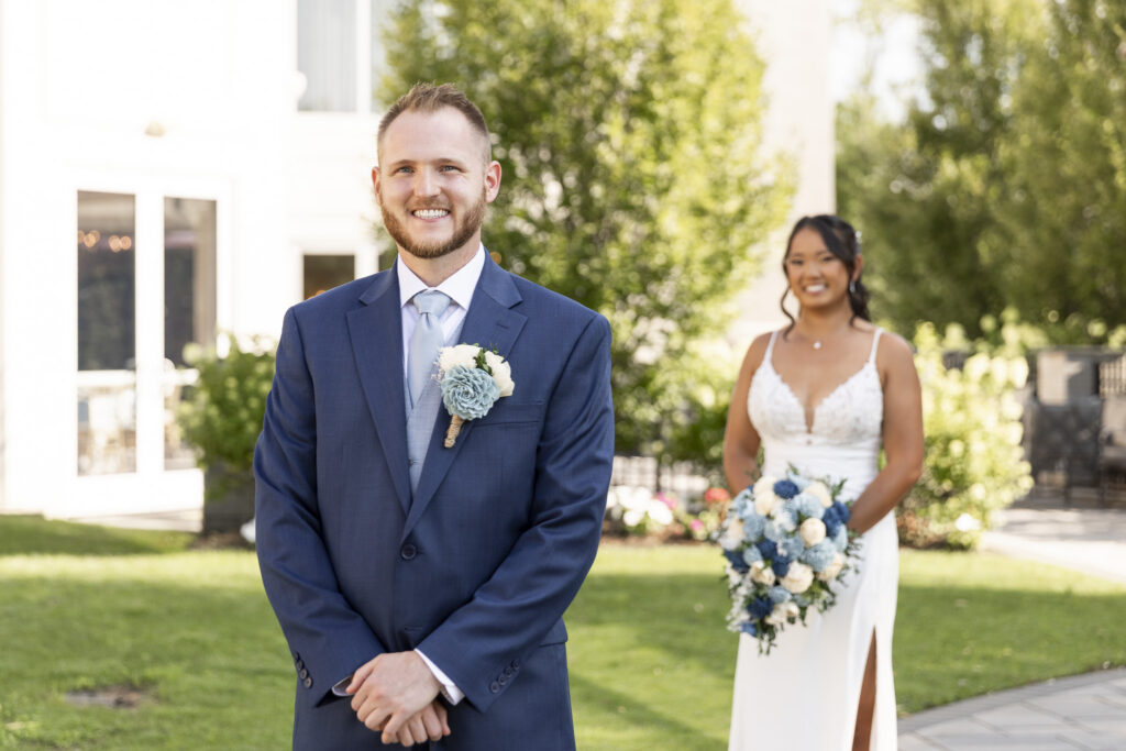 Groom smiles while waiting for his bride during a first look in the sunlit gardens at The Mansion on Main Street, with the bride approaching behind him carrying a blue and white bouquet.