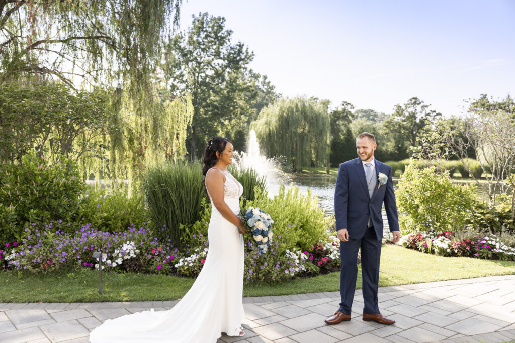 Bride and groom see each other during their first look beside colorful gardens and a fountain at The Mansion on Main Street, surrounded by weeping willow trees in the afternoon light.