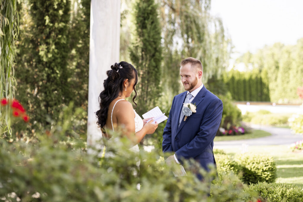 Bride reads private vows from a small notebook to the groom in the gardens at The Mansion on Main Street, framed by greenery and soft foreground foliage.