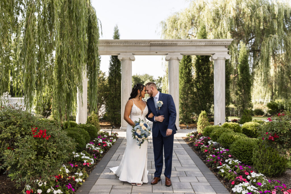 Newlyweds walk arm in arm along a garden path at The Mansion on Main Street, touching foreheads beneath a columned structure surrounded by weeping willows and colorful flowers.