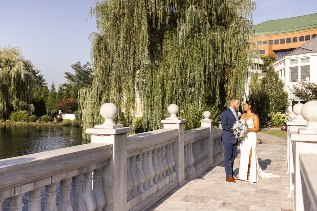 Bride and groom stand together on a stone bridge overlooking the pond and weeping willow trees during their wedding at The Mansion on Main Street.