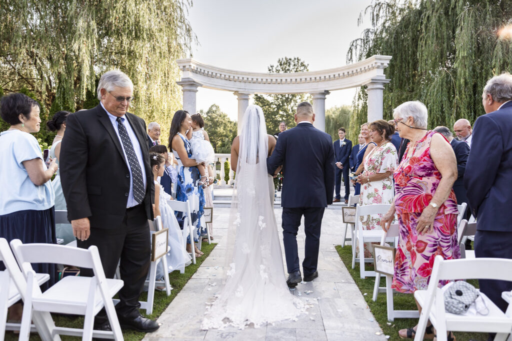 Bride walks down the outdoor ceremony aisle at The Mansion on Main Street with an escort, her long lace-edged veil trailing behind her as guests stand on either side.