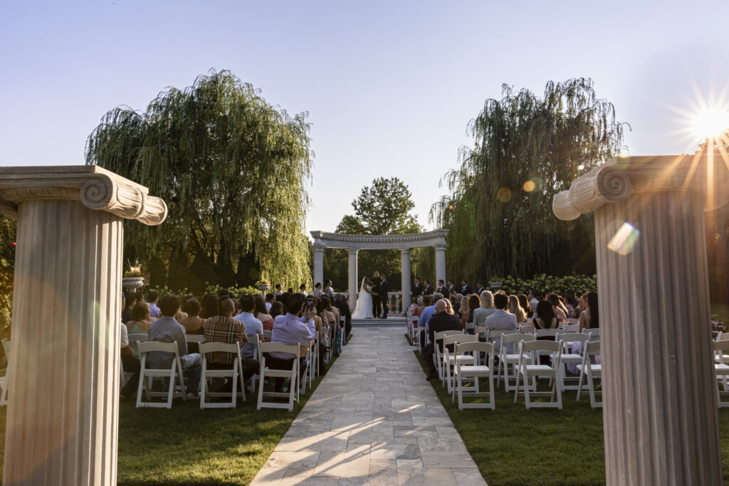 Wide view of an outdoor wedding ceremony at The Mansion on Main Street, with guests seated along a stone aisle leading to the couple beneath a curved columned arbor as the evening sun shines through the trees.