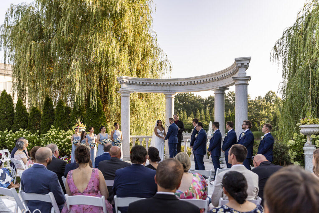 Bride and groom stand hand in hand beneath the curved stone arbor during their outdoor ceremony at The Mansion on Main Street, surrounded by their wedding party, seated guests, weeping willow trees and lush gardens.