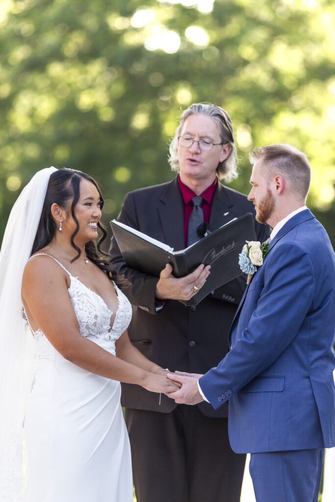 Bride and groom hold hands and smile at one another while exchanging vows during their outdoor south jersey wedding ceremony, with their officiant standing behind them.
