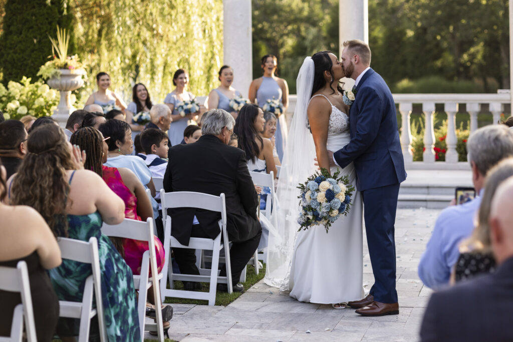 Bride and groom share a kiss at the end of their outdoor south jersey wedding ceremony as seated guests applaud and their wedding party celebrates behind them.