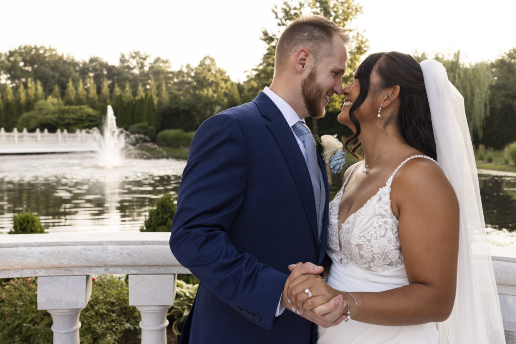 Newlyweds smile with their foreheads touching while holding hands beside the pond and fountain in the gardens of The Mansion on Main Street.