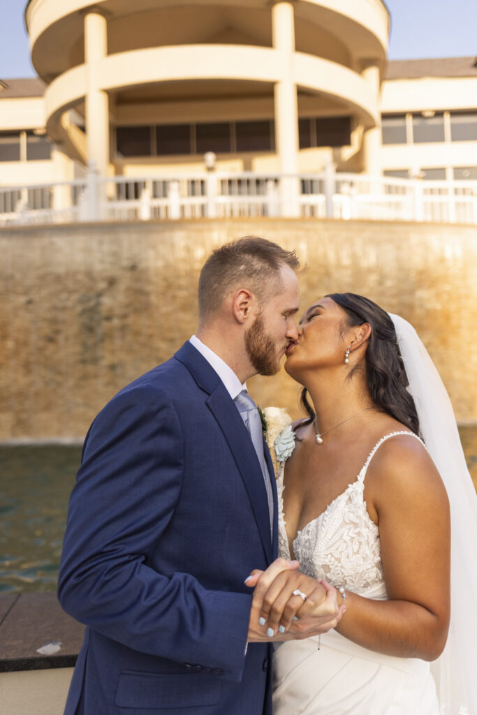 Close portrait of the newlyweds kissing beside the water during golden hour, with the architecture of The Mansion on Main Street softly illuminated behind them.