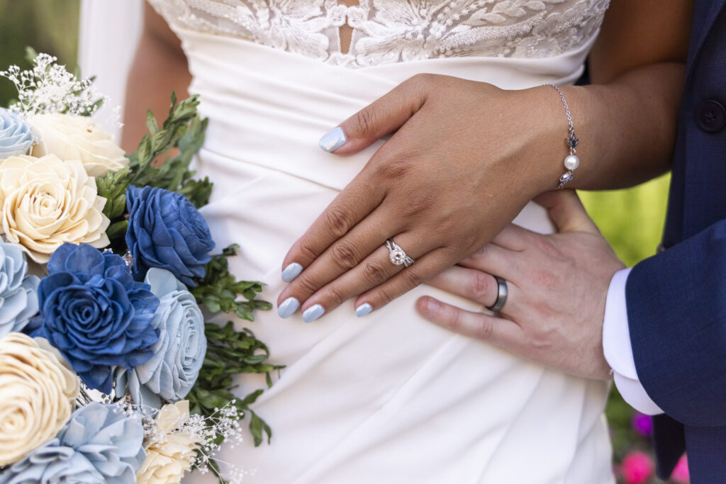 Close-up of the newlyweds’ hands displaying their wedding rings beside a bouquet of blue and ivory flowers during their south jersey wedding portraits.