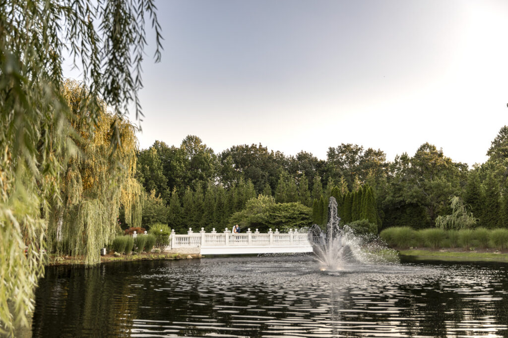 Wide view across a reflective pond and fountain toward a white bridge, where the newlyweds stand together surrounded by lush gardens during their south jersey wedding.