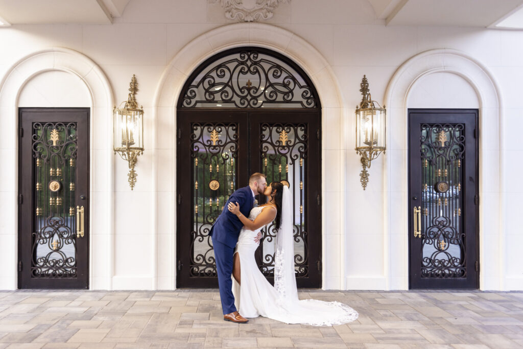 Bride and groom share a kiss in front of the grand wrought-iron doors and arched architecture at The Mansion on Main Street, with her long lace-edged veil trailing across the stone patio.