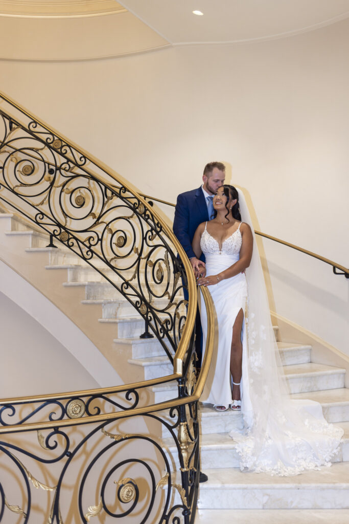 Groom kisses the bride's forehead as they pose together on the sweeping marble staircase at The Mansion on Main Street, framed by ornate black and gold wrought-iron railings.