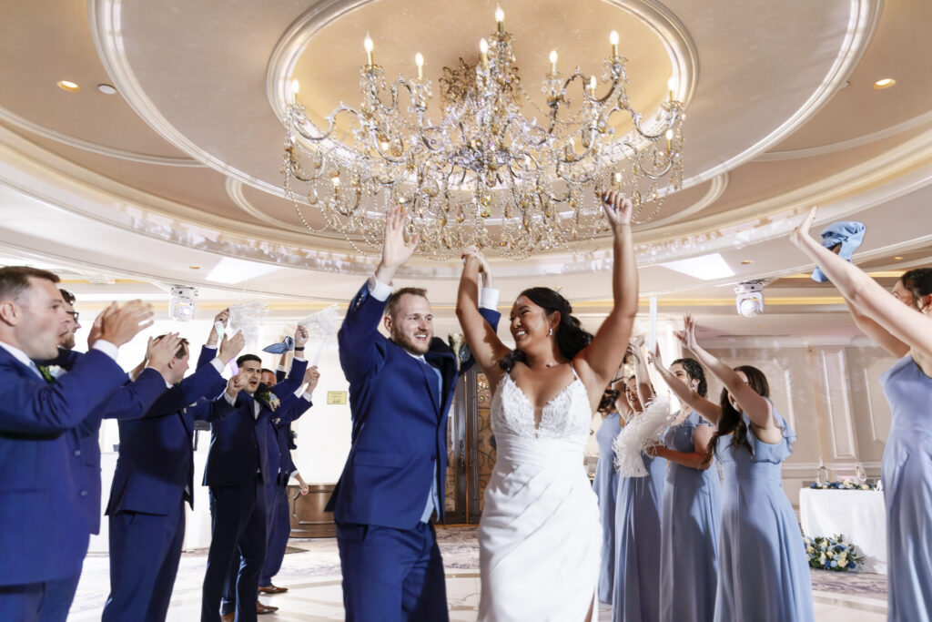 Newlyweds enter their reception hand in hand with their arms raised as the wedding party cheers beneath a large crystal chandelier at The Mansion on Main Street.