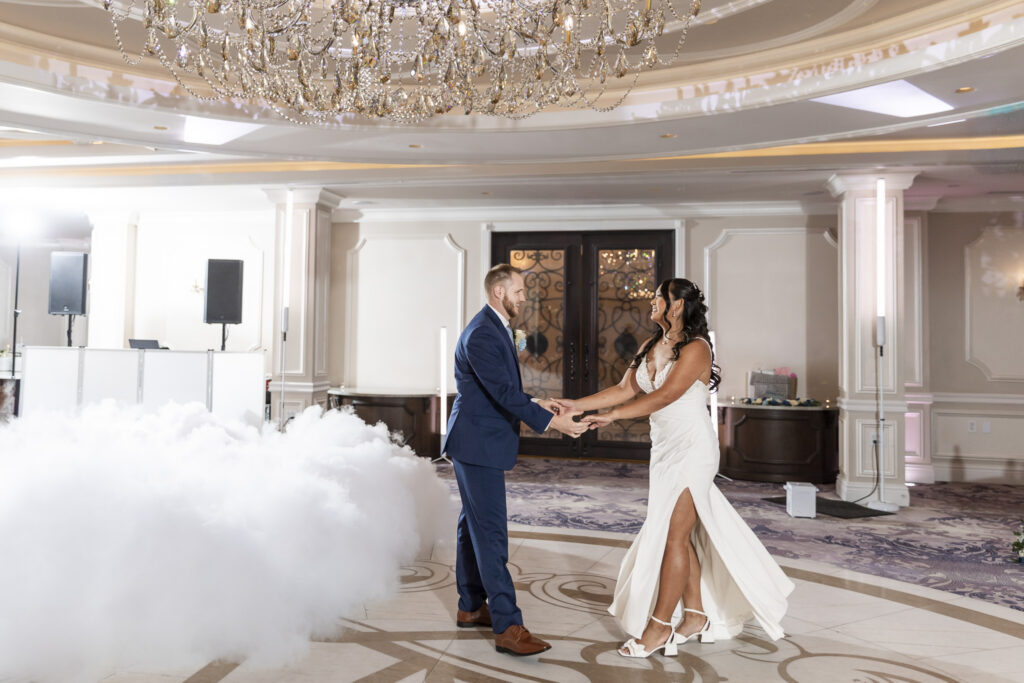 Newlyweds hold hands during their first dance at The Mansion on Main Street, with low-lying fog drifting across the ballroom floor beneath a large crystal chandelier.