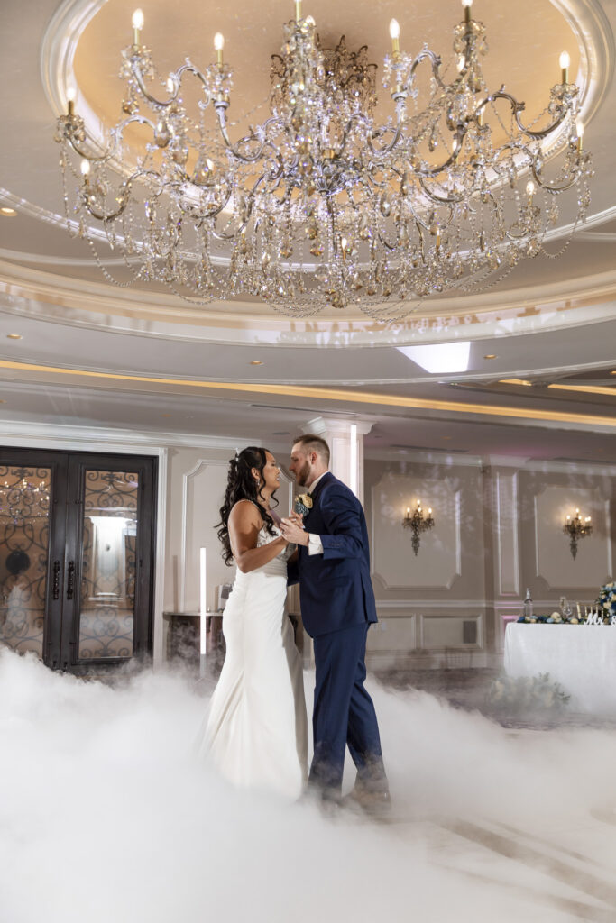 Newlyweds share a slow dance beneath an elaborate crystal chandelier, surrounded by low-lying fog that blankets the ballroom floor during their wedding reception.