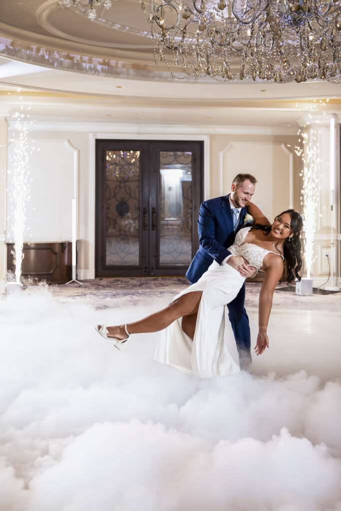 Groom dips the bride during their first dance as indoor sparklers glow on either side and low-lying fog covers the ballroom floor at The Mansion on Main Street.