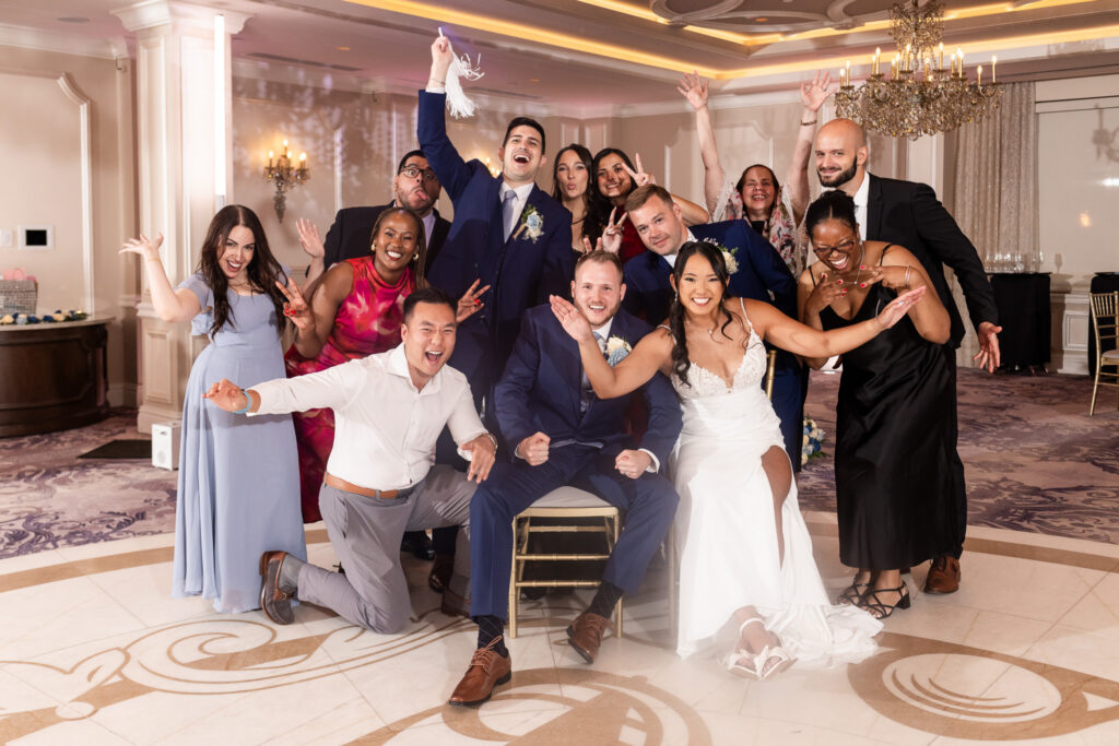Bride and groom sit together in the center of a lively group of friends and family, who laugh, wave and pose around them during their south jersey wedding reception.