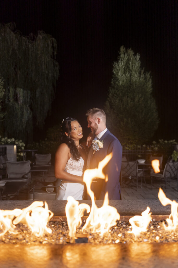 Newlyweds stand close together outdoors at night behind a glowing fire feature, smiling at each other during an evening portrait at their south jersey wedding.