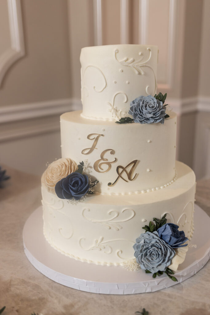 Three-tier white wedding cake decorated with delicate piping, blue and ivory flowers, greenery, and gold initials sits on display during an elegant south jersey wedding reception.