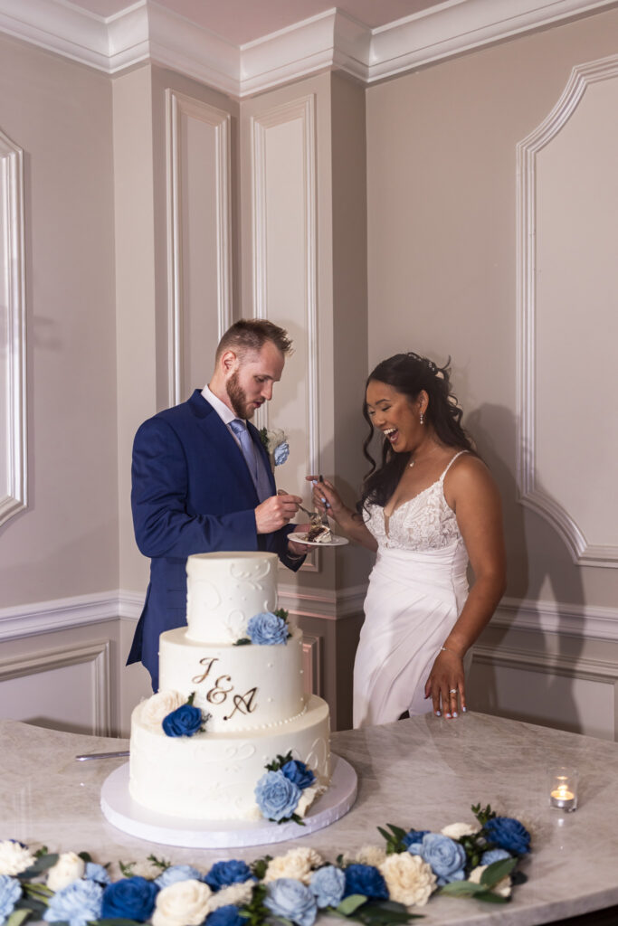 Bride laughs while sharing wedding cake with the groom beside a three-tier white cake decorated with blue and white flowers during their reception at The Mansion on Main Street.
