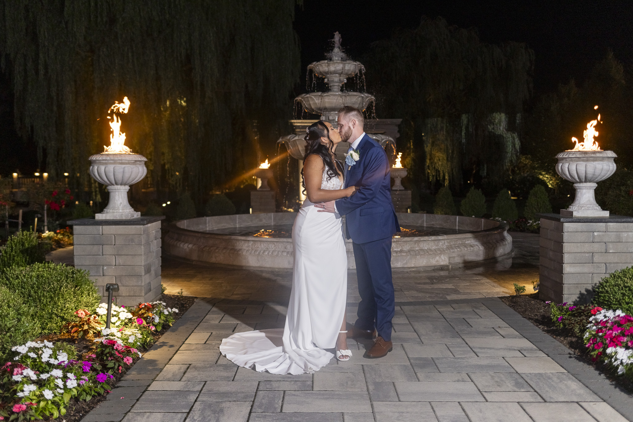 Bride and groom kiss in the illuminated gardens at night, framed by glowing fire features and a tiered stone fountain at The Mansion on Main Street.