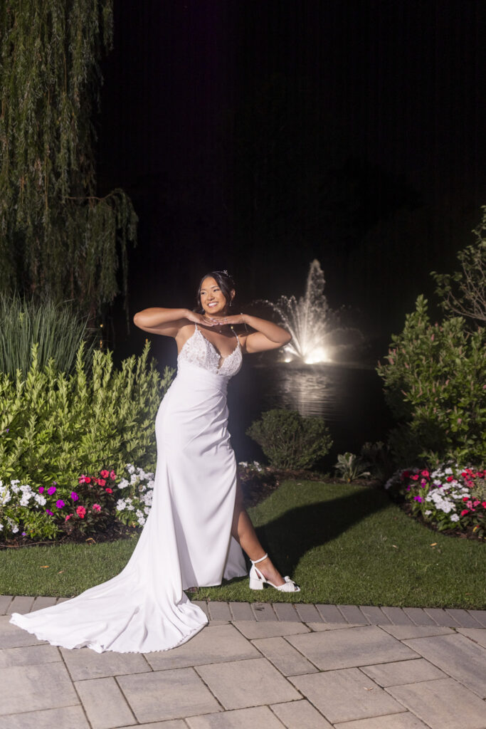 Bride poses in her lace and satin wedding gown beside colorful flower beds, with an illuminated fountain reflecting across the pond behind her at The Mansion on Main Street.