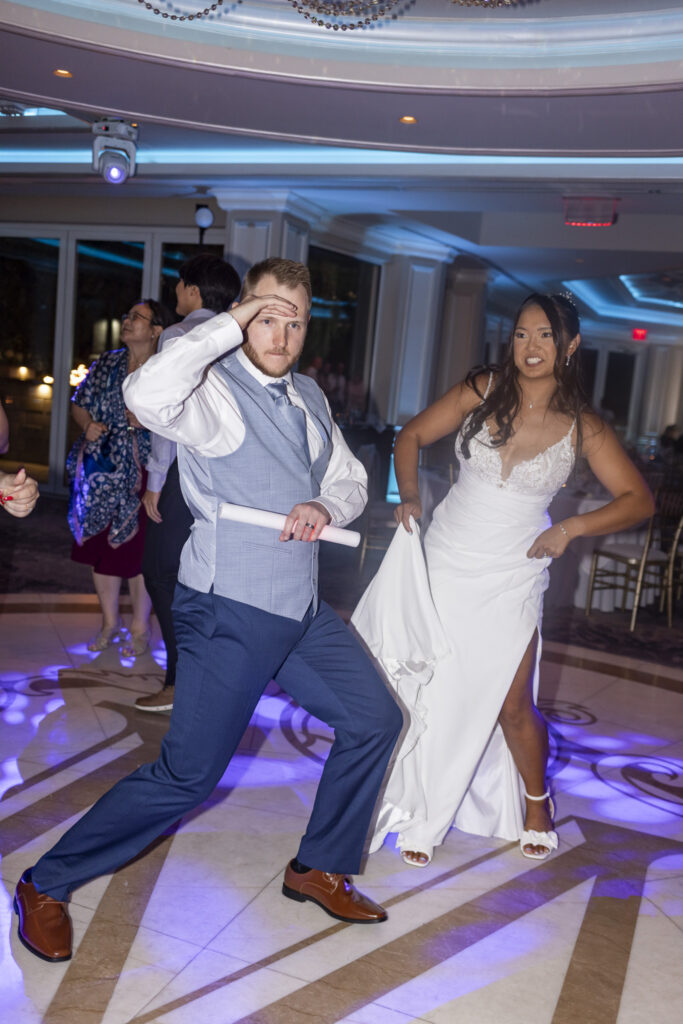 Groom strikes a playful pose with a glowing foam light stick on the dance floor while the bride dances behind him, lifting the skirt of her wedding dress amid colorful reception lighting.