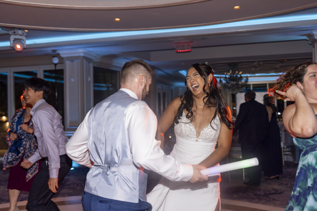 Bride laughs while dancing with the groom during their wedding reception, surrounded by guests beneath blue ballroom lighting as the groom holds a glowing foam light stick.