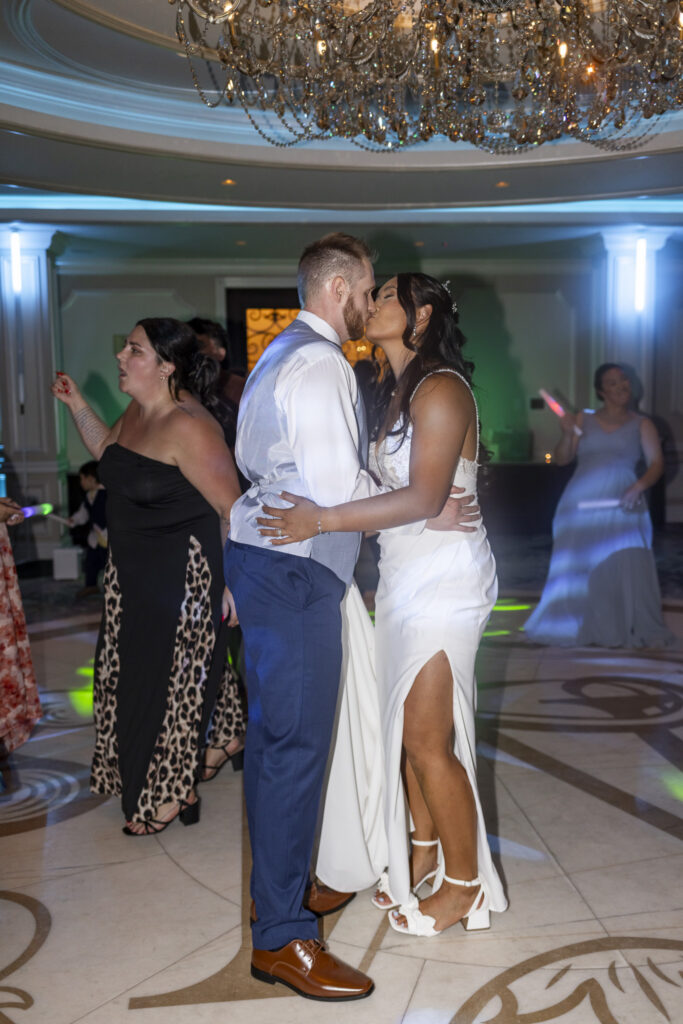 Bride and groom kiss on the dance floor during their reception at The Mansion on Main Street, surrounded by guests, colorful DJ lighting and a sparkling crystal chandelier overhead.